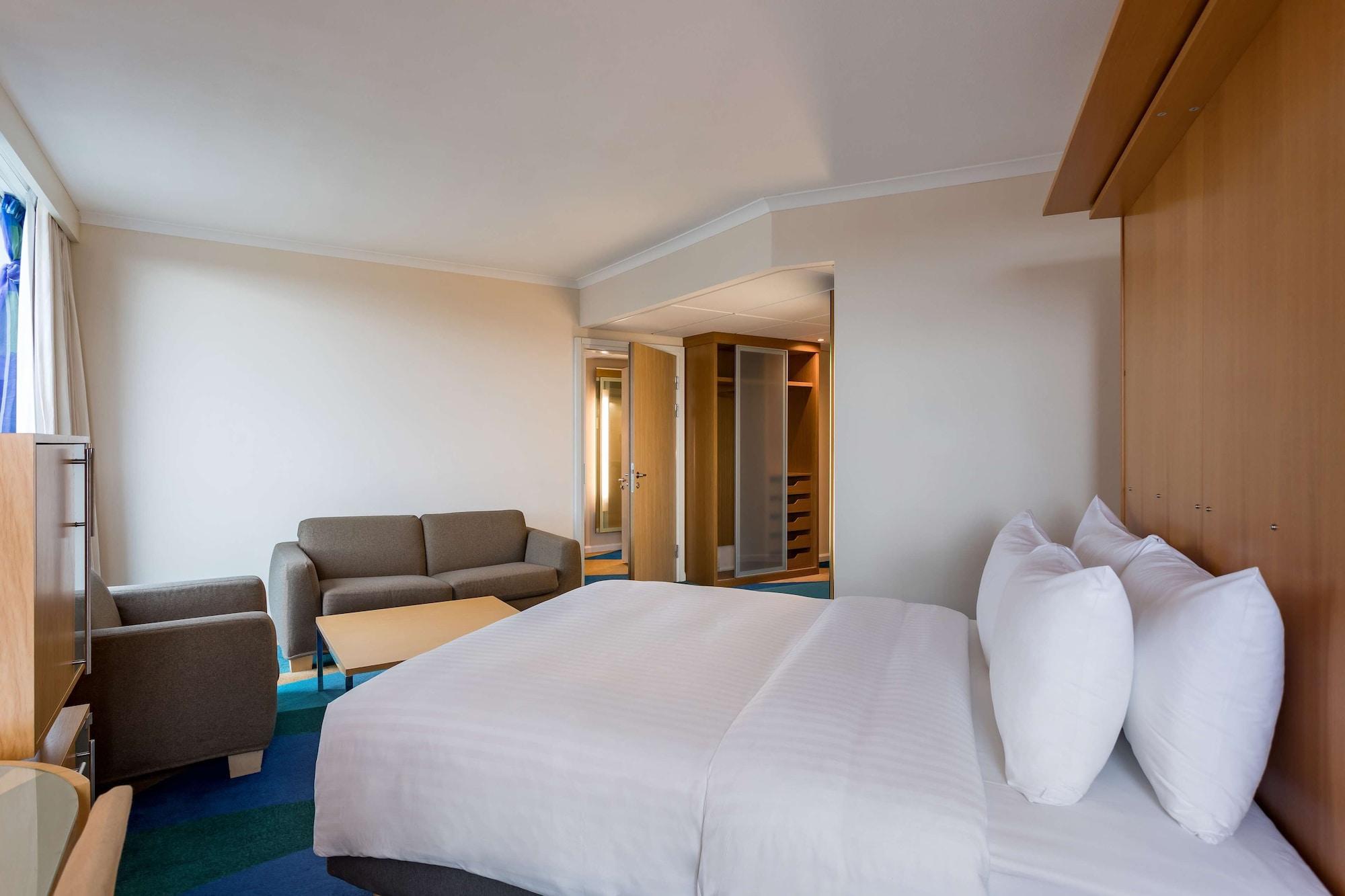 Radisson Blu Hotel London Stansted Airport Stansted Mountfitchet Esterno foto The photo shows a modern hotel room featuring a large bed with two white pillows at the forefront. To the left, there is a small seating area with a gray sofa and a coffee table. The room has a light-colored, minimalist design with neutral walls and 