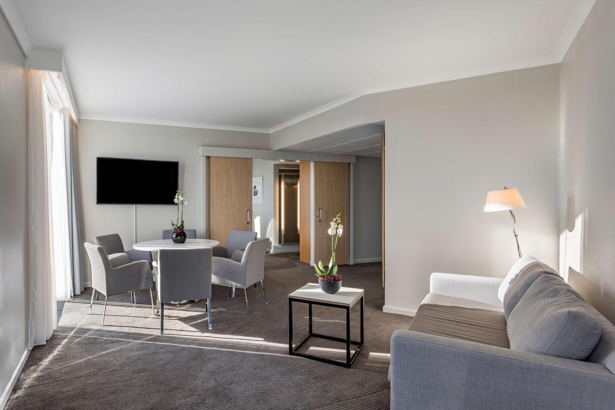 Radisson Blu Hotel London Stansted Airport Stansted Mountfitchet Esterno foto The image depicts a modern hotel room or suite. It features a comfortable living area with a light gray sofa and a small coffee table. There are two chairs around a circular table, suggesting a space for dining or conversation. A flat-screen televisi